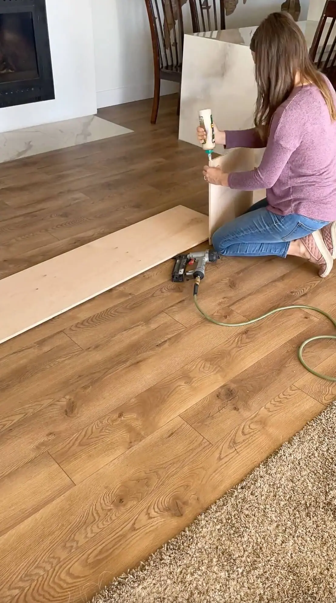 A woman assembling a bench on a newly installed wood-look floor. She is applying wood glue to the joints before securing them with a nail gun, working in a bright living space with a fireplace in the background.